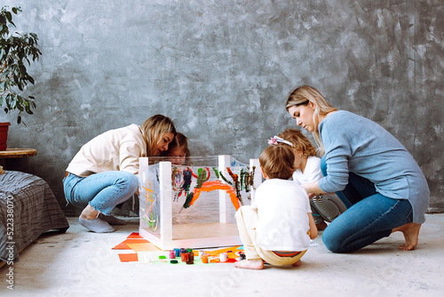 Photo Attentive little girls sitting on haunches on art lesson, painting with mothers teachers with brushes on stretch film