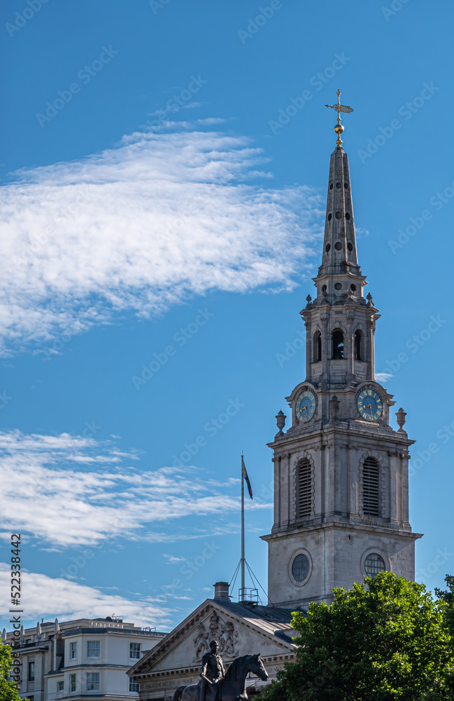 London, UK- July 4, 2022: Trafalgar Square. Spire and pediment of Saint Martin-in-the-fields church against blue cloudscape and green foliage at bottom. King George IV statue.