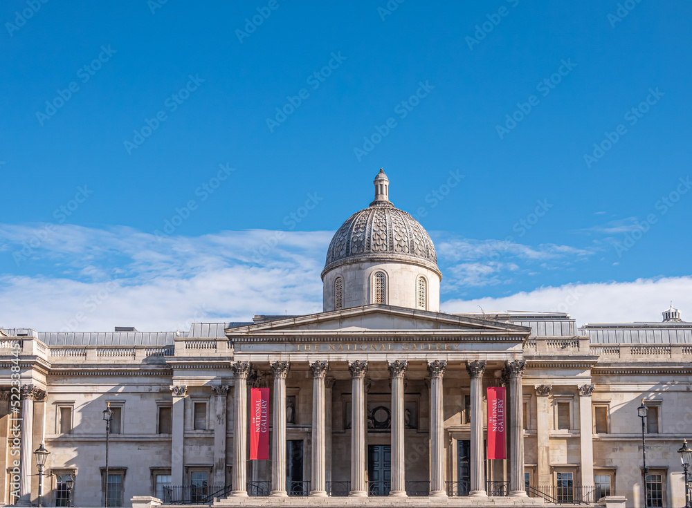 London, UK- July 4, 2022: Trafalgar Square. Central part with dome and pediment of National Gallery building under blue cloudscape. Red banners add color.