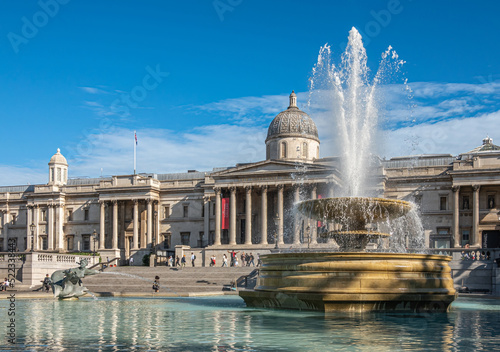 London, UK- July 4, 2022: Trafalgar Square. Wide view on Full blowing water Jellycoe fountain with National Gallery building in back under blue cloudscape.