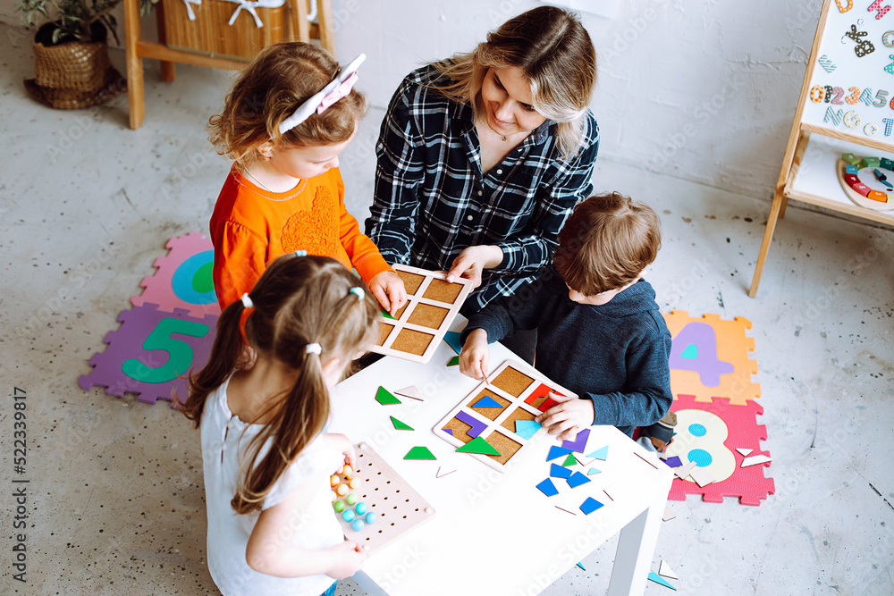 Top view of group of preschoolers. Young blond woman helping pupils to play board games, collecting geometric figures. - obrazy, fototapety, plakaty 