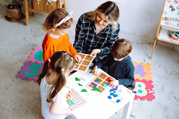 Top view of group of preschoolers. Young blond woman helping pupils to play board games, collecting geometric figures.