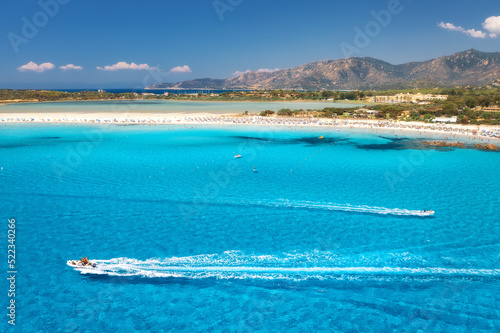 Speed boat in transparent blue water and white sandy beach at sunny day in summer. Travel in Sardinia, Italy. Top view from drone of sea coast, motorboats, mountain, trees, sky. Tropical. Aerial view