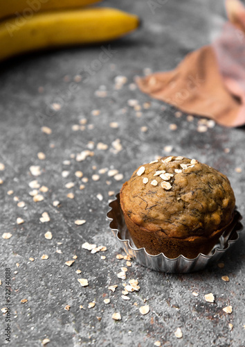 Vertical photograph of a fresh baked banana muffin, with bananas in background and sprinkled with oats