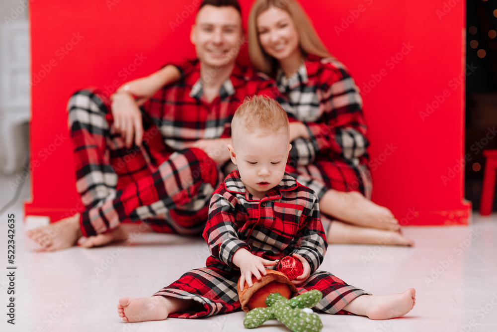 Cute baby boy playing while parents relaxing behind at home together, smiling active boy entertaining with toy cactus on floor, happy family spending time together on weekend on red wall background.
