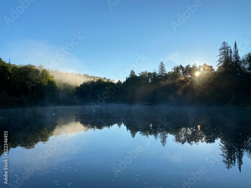 Paysage calme d'une forêt et de l'eau d'une rivière à l'aube. Reflet d'une montagne sur un lac au petit matin. Lever de soleil avec brume sur l'eau. Rayons de soleil au travers le brouillard.