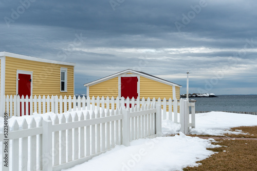 The exterior of colorful wooden sheds with a red door overlooking the blue ocean and cloudy sky. There's a white picket fence in the yard with white snow piled up next to the beach hut style building.