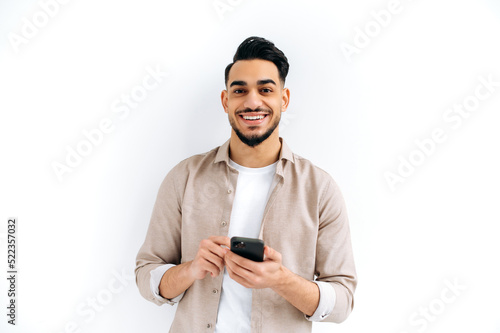 Handsome positive Arabian or Indian modern guy, holding smartphone in his hands, messaging, looking excitedly at the camera, standing over isolated white background, smiling © Kateryna
