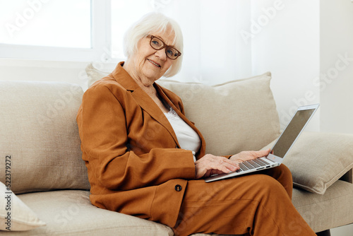 a pleasant, sweetly smiling elderly old lady is sitting with a laptop on her lap on a beige sofa dressed in a stylish brown suit and looking at the camera © Tatiana