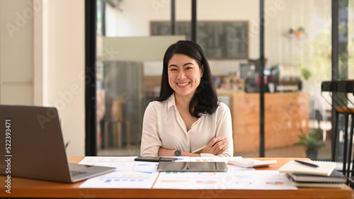 Confident asian woman employee sitting with arms crossed at her workplace and smiling to camera © Prathankarnpap
