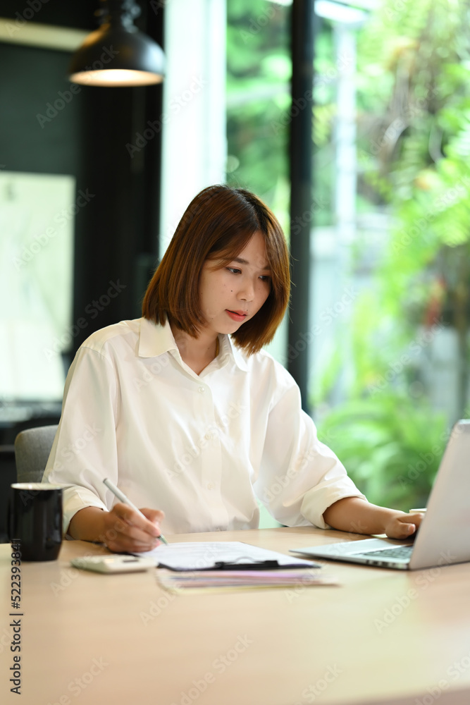 Concentrated female economist using laptop an working with document at office desk Concentrated female economist using laptop an working with document at office desk