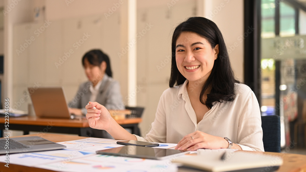 Happy asian female office worker sitting in corporate office ad smiling to camera Happy asian female office worker sitting in corporate office ad smiling to camera
