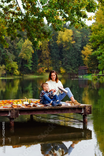 A woman and a child drink tea from a thermos in autumn on a pier by the lake. Mother and son spend time together in nature.