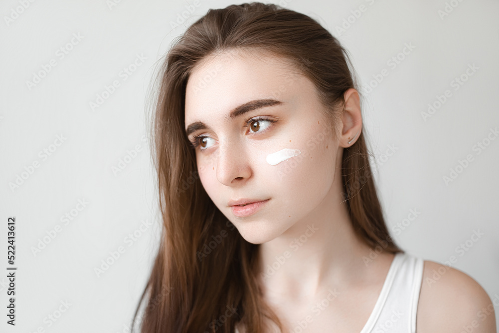 A young beautiful girl uses a cream to take care of her facial skin. Home care procedures for women. Face without wrinkles close-up. Brunette woman on gray background A young beautiful girl uses a cream to take care of her facial skin. Home care procedures for women. Face without wrinkles close-up. Brunette woman on gray background