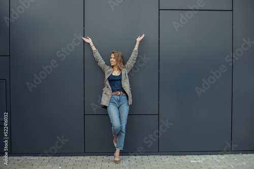 Fototapeta Naklejka Na Ścianę i Meble -  Full length photo of happy young businesswoman standing on one leg leaning on gray wall of office building with raised arms. Urban lifestyle, entrepreneur, well dressed lady, successful people