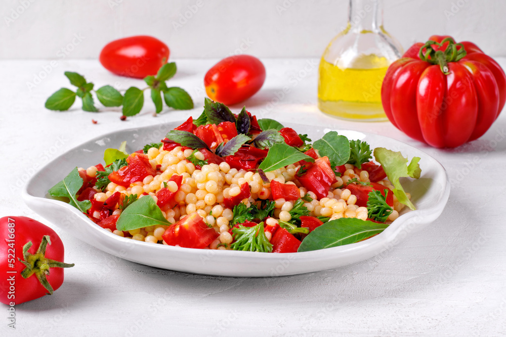 Warm salad with ptitim pasta, grilled peppers, tomato, arugula, basil and olive oil served on the white plate Warm salad with ptitim pasta, grilled peppers, tomato, arugula, basil and olive oil served on the white plate