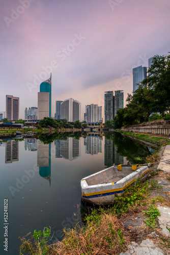 Urban landscape of Jakarta, Indonesia, featuring high-rise buildings, river reflection, and a traditional wooden boat under a purple twilight sky.