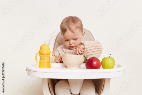 Portrait of concentrated baby wearing beige sweater sitting in a child's chair, isolated on a white background, holding spoon in hand and eating porridge with attentive expression.