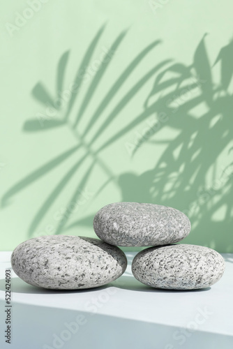 A podium made of stones on a gray table against a green wall with a shadow from the leaves of a palm tree. Showcase for product promotion, beauty, natural cosmetic