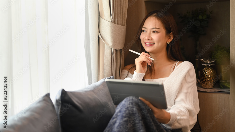Satisfied woman holding tablet and looking through the window, spending leisure time at home in autumn day Satisfied woman holding tablet and looking through the window, spending leisure time at home in autumn day