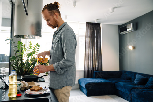 Young ginger man drinking lunch while making lunch in kitchen