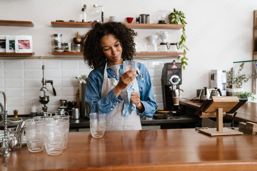 Black barista woman wearing apron wiping glasses while working in cafe Black barista woman wearing apron wiping glasses while working in cafe