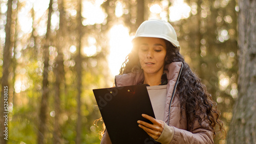 Millennial female technician ecologist looking up at treetops, Young indian woman in hardhat with clipboard taking measures checking trees. Forestry engineer in park. Supervising wildlife sanctuary photo