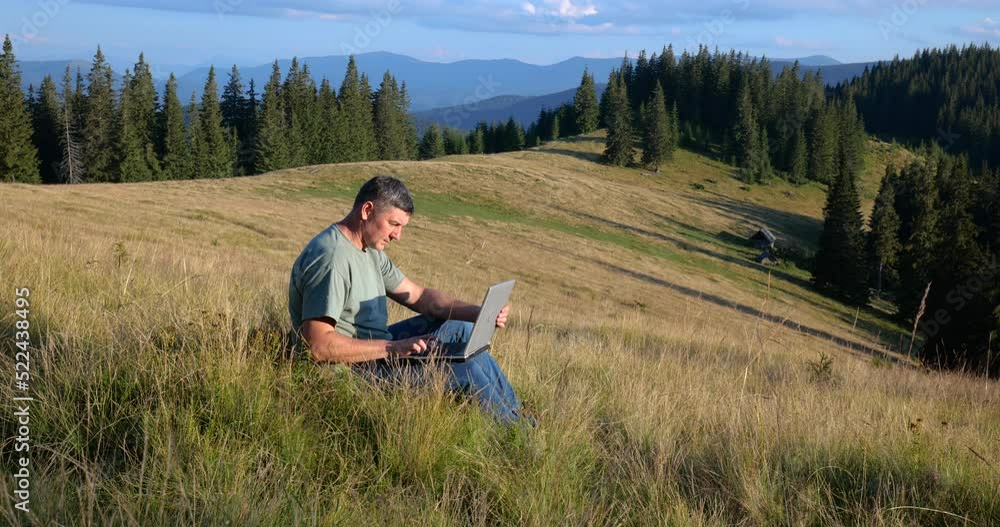A man sits on a beautiful meadow in the mountains, works on a laptop A man sits on a beautiful meadow in the mountains, works on a laptop