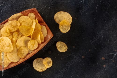 Potato chips or crisps, a salty snack in a wooden bowl, overhead flat lay shot on a black slate background with copy space