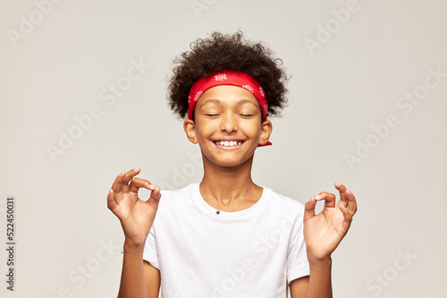 Funny laughing kid in white mockup shirt with copy space for your text pretending doing yoga practice keeping fingers in mudra sign wearing red bandana on head with afro hairstyle on gray background