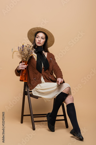 Stylish woman in sun hat holding plants in vase on beige background