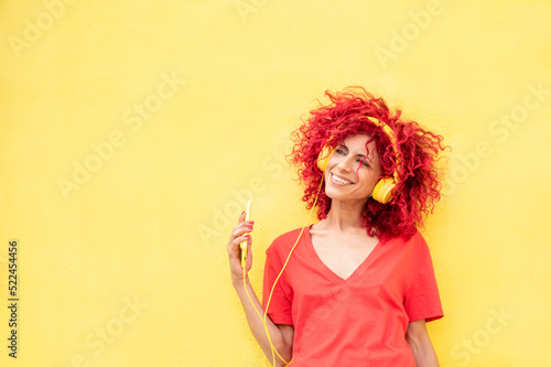 beautiful woman with a red afro hair holding a smartphone and listen to the music with a yellow headphones over yellow background
