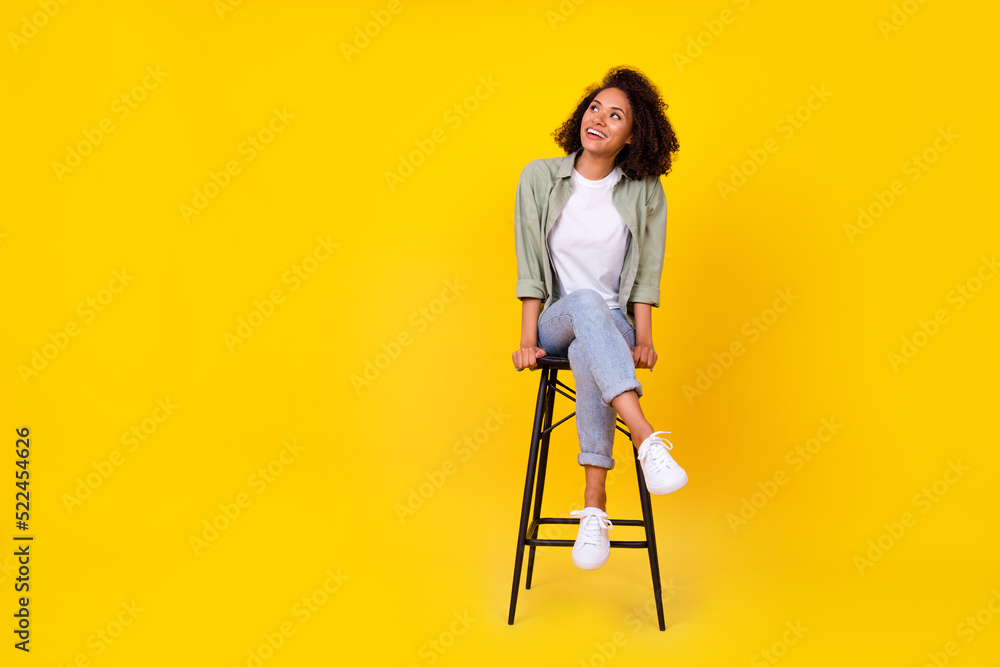 Full length photo of cute young lady wavy hair sit stool look empty space dressed stylish khaki garment isolated on yellow color background
