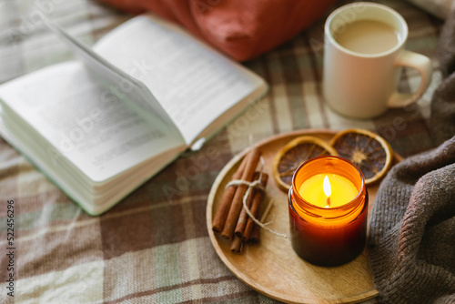 Burning candle with wooden wich in amber glass gar, open book and cup of coffee or tea. Autumn home decor. Cozy fall mood.