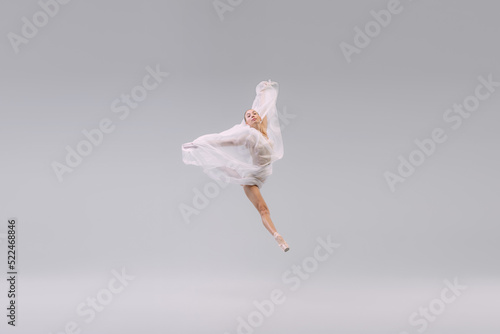Fototapeta Naklejka Na Ścianę i Meble -  Portrait of young ballerina dancing, jumping with transparent fabric isolated over grey studio background. Leap