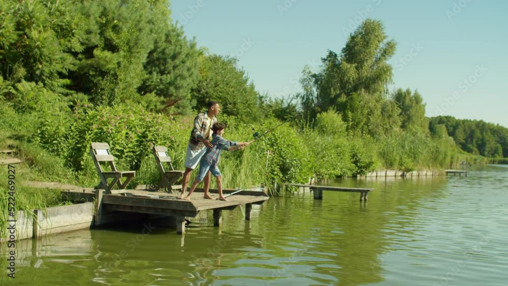Loving black father teaching lovely school age multicultural son to cast spin fishing rod from wooden pier on lake, showing support and care while multiethnic family enjoying angling on summer day. Loving black father teaching lovely school age multicultural son to cast spin fishing rod from wooden pier on lake, showing support and care while multiethnic family enjoying angling on summer day.