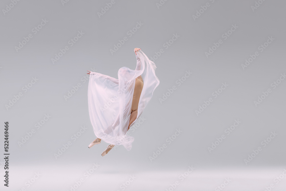 Naklejka premium Portrait of young ballerina dancing, performing in beige cloth, covered with fabric isolated over grey studio background