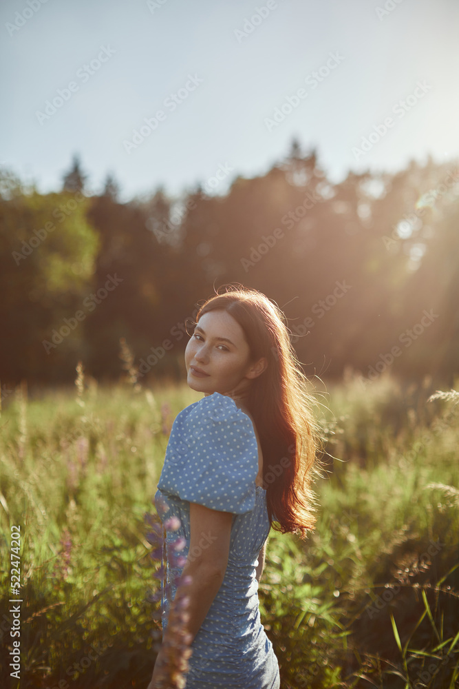 Side view of a charming young woman in a blue dress walks in a field. The girl is enjoying the good weather and sun.