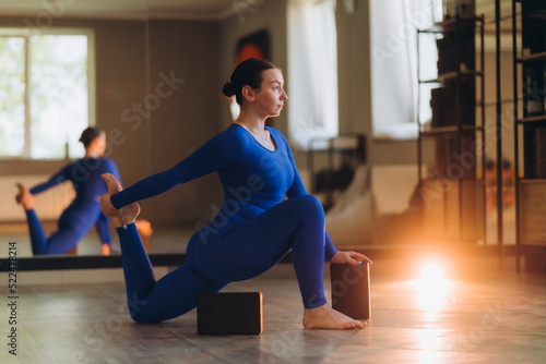Portrait of a beautiful young European curvy woman doing sports in the studio. A girl in leggings and a blue top stands on one column and stretches. A look to the side. Yoga, fitness, sports.