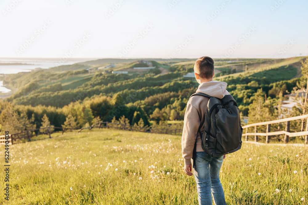 Tourist with backpack standing on top of hill in grass field and enjoying beautiful landscape view. Rear view of teenage boy hiker resting in nature. Active lifestyle. Concept of local travel