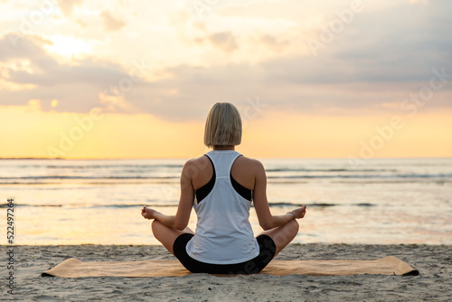 yoga, mindfulness and meditation concept - woman meditating in lotus pose on beach over sunset