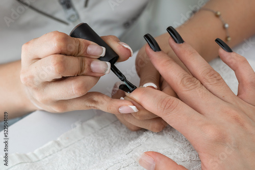 close-up detail shot of a manicurist s hands painting the nails with a black nail polish. designing and beautifying the nails of a woman in the beauty salon. varnishing the nails. dark style.