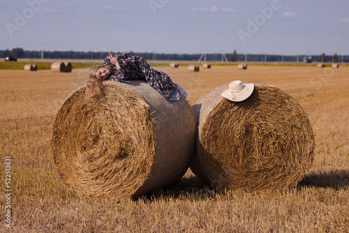 A blonde girl with long hair in a white hat is resting and posing near the sheaves of hay in a field on a sunny day
