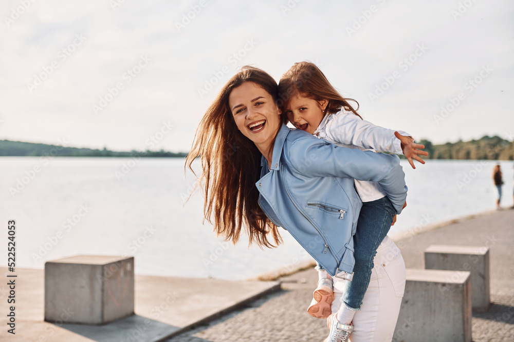 Embracing each other and smiling. Young mother with her daughter having fun outdoors near the lake at summer