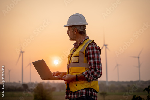 Portrait of engineer wearing yellow vest and white helmet using a computer laptop on site at wind turbines field or farm, Sustainable energy concept