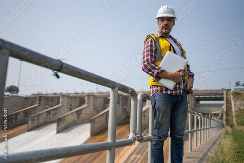 Portrait of engineer wearing yellow vest and white helmet with laptop computer Working day on a water dam with a hydroelectric power plant. Renewable energy systems, Sustainable energy concept