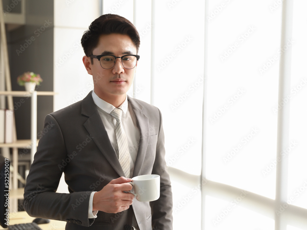 A young Asian businessman wearing a fit suit, wearing eyeglasses, has the personality of a successful man. Holding cup of coffee in office in morning sunshines. A young Asian businessman wearing a fit suit, wearing eyeglasses, has the personality of a successful man. Holding cup of coffee in office in morning sunshines.