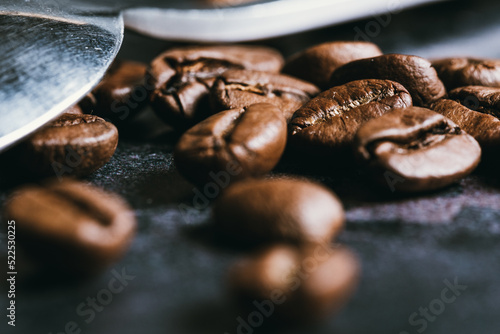 Close up of some Toasted Coffee Beans near to a Scratched Metal Spoon over a Black Concrete Texture Backdrop. Coffee concept