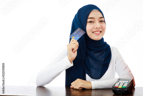 A beautiful young Muslim woman with cheerful smile, friendly personality, and self-confidence holding credit card sitting next to credit card reader.