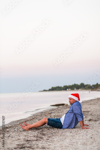 Vertical view of beach vacation travel. Vacation concept. Christmas holiday. Young man sitting in a Santa hat on paradise beach island on the seaside background with a bottle of whiskey in hands. 